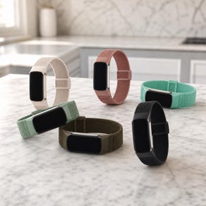 May include: Six fitness trackers with rectangular black screens and metallic frames, each featuring a woven fabric band in a different color: beige, dusty rose, mint green, olive green, black, and a lighter sage green, arranged on a surface.