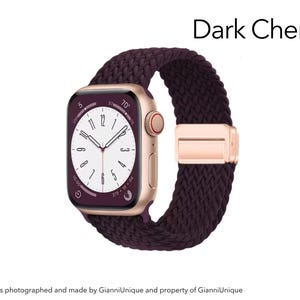 May include: A smartwatch with a gold-tone case and a dark cherry braided solo loop band. The watch face displays a white dial with black numerals and hands, showing the text Dark Cherry. The band features a metallic gold-tone adjustable clasp.