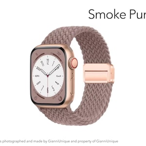 May include: A smartwatch with a metallic rose gold case and a smoke purple braided solo loop band. The watch face displays a white dial with black numerals and hands. The text Smoke Purple is visible in the upper right corner of the image.