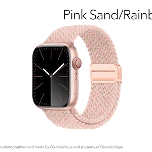 May include: A smartwatch with a rose gold case and a black screen featuring white clock hands. The watch band is a braided pink sand material with subtle rainbow-colored flecks. The text Pink Sand/Rainbow is displayed above the watch.