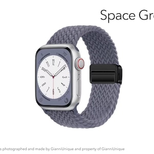 May include: A smartwatch with a rectangular silver case and a white analog clock face featuring black numerals. The watch is attached to a textured, braided grey strap secured by a black metal clasp. The text Space Grey appears above the watch.