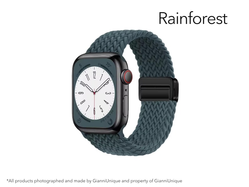 May include: A smartwatch with a dark metallic rectangular case and a dark teal braided solo loop band. The watch face displays a white dial with black numerals and hands. The word Rainforest is printed in black text above the watch.