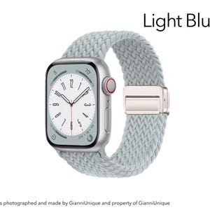 May include: A silver smartwatch with a light blue braided solo loop band. The watch face displays a white dial with black numerals and hands. The text Light Blue is visible in the upper right corner. The side features a digital crown with a red ring.