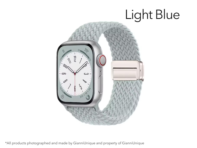 May include: A silver smartwatch with a light blue braided solo loop band. The watch face displays a white dial with black numerals and hands. The text Light Blue is visible in the upper right corner. The side features a digital crown with a red ring.