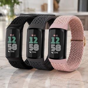 May include: Three fitness trackers with digital displays showing a heart icon, the time 12:58, and 7,546 steps. The trackers feature braided elastic bands in black, dark gray, and light pink, each with a small metallic adjustment buckle on the side.