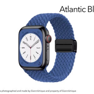 May include: A smartwatch with a black rectangular case and a textured Atlantic Blue braided band. The watch face features a white dial with black numerals and a red-accented crown. The text Atlantic Blue is displayed in the upper right corner.