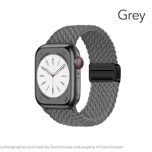May include: A smartwatch with a dark grey rectangular case and a grey braided elastic watch band. The watch face displays a white background with black numerals and hands. A black magnetic clasp secures the band. The word Grey is visible.