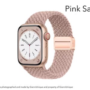 May include: A smartwatch with a rectangular gold-tone case and a pink sand braided band. The watch face displays a white dial with black numerals and hands. The text Pink Sand is visible in the upper right corner of the image.