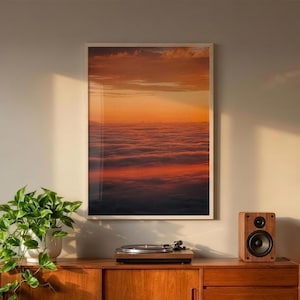 Könnte beinhalten: Ein rechteckiger gerahmter Wandkunstdruck mit einer Ansicht aus großer Höhe auf eine dichte Wolkenschicht bei Sonnenuntergang, die lebendige orange, rote und tiefblaue Verläufe zeigt. Der Druck befindet sich in einem dünnen weißen Rahmen.