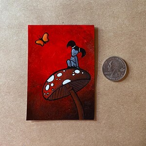 May include: A small painting featuring a whimsical scene. A blue figure sits atop a mushroom with white spots against a red background. An orange butterfly flies nearby. A U.S. quarter is included for size reference.