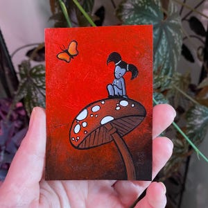 May include: A small, rectangular painting with a red background features a whimsical scene. A gray figure with black hair sits atop a brown mushroom with white spots. An orange butterfly flutters nearby. The artwork has a fantasy theme.