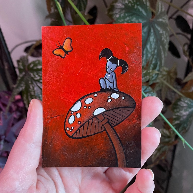 May include: A small, rectangular painting with a red background features a whimsical scene. A gray figure with black hair sits atop a brown mushroom with white spots. An orange butterfly flutters nearby. The artwork has a fantasy theme.