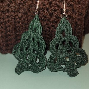 May include: A pair of green crocheted earrings shaped like Christmas trees. The earrings are hanging from silver hooks.