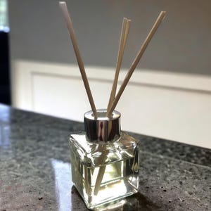 Può includere: Un flacone diffusore quadrato in vetro trasparente con tappo argentato e diversi bastoncini di legno. Il flacone contiene un liquido giallo chiaro. Il diffusore è appoggiato su un piano di lavoro grigio maculato, suggerendo un prodotto per la profumazione della casa.
