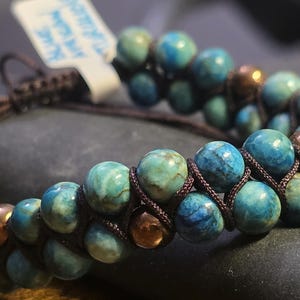 May include: A handcrafted bracelet with blue and green gemstone beads, interspersed with copper-toned beads. The beads are strung on a dark brown cord, forming a double-strand design. A small tag is attached to the bracelet.