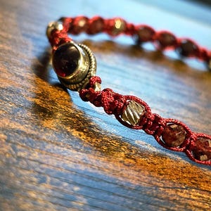 Puede incluir: Primer plano de una pulsera de macramé roja con una gema grande, redonda y de color rojo oscuro y cuentas más pequeñas, facetadas, rojas y doradas. La pulsera está sobre una superficie de madera, destacando el intrincado anudado y el trabajo de cuentas.