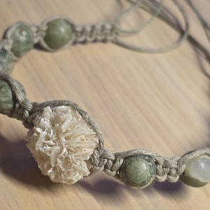 May include: Handmade macrame bracelet featuring olive green gemstone beads and a central cluster of cream-colored crystals. The bracelet is crafted with a natural-toned cord, showcasing a bohemian style.