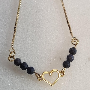 Puede incluir: Un collar dorado con un colgante en forma de corazón y cuentas azul oscuro. El collar tiene una cadena delicada y está diseñado para usarse alrededor del cuello. El colgante de corazón es abierto y las cuentas están espaciadas uniformemente.