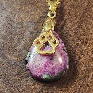 May include: A gold chain necklace with a pink and green gemstone pendant featuring a gold paw print charm.