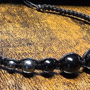 May include: A black beaded necklace with a macrame cord. The necklace features a series of black and silver beads of varying sizes. The beads are strung together with a dark gray macrame cord.