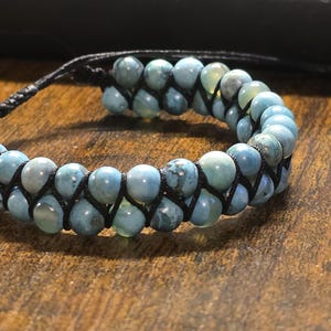 May include: A blue beaded bracelet with a woven design. The bracelet features light blue and gray beads strung together with black cord. The bracelet is adjustable and rests on a wooden surface.