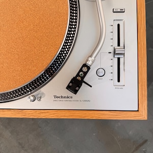 Puede incluir: Un tocadiscos de tracción directa Technics SL-1200GR2 con placa superior plateada, alfombrilla de corcho y marco de madera. El dispositivo cuenta con un brazo plateado, un control deslizante de ajuste de tono con botón de reinicio y marca.