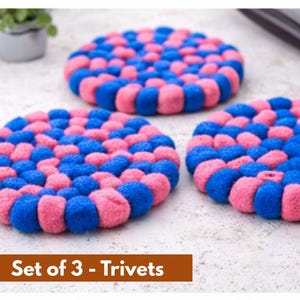 May include: Set of three round trivets in a vibrant pattern of blue and pink. The trivets are made of a soft, textured material and are arranged on a light-colored surface. The text "Set of 3 - Trivets" is at the bottom.