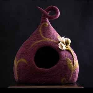 May include: A dome-shaped felted pet cave in a deep plum color with a curled top point. The structure features a circular opening and is decorated with embroidered olive green vine patterns and three-dimensional white felted flower blossoms.