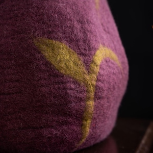 Peut inclure: Un objet en laine feutr&eacute;e magenta fonc&eacute; &agrave; la surface textur&eacute;e orn&eacute; d'un motif en forme de feuille jaune moutarde. Le coin sup&eacute;rieur droit affiche un logo avec le texte The Oak Studio en fine police dor&eacute;e sur fond sombre.