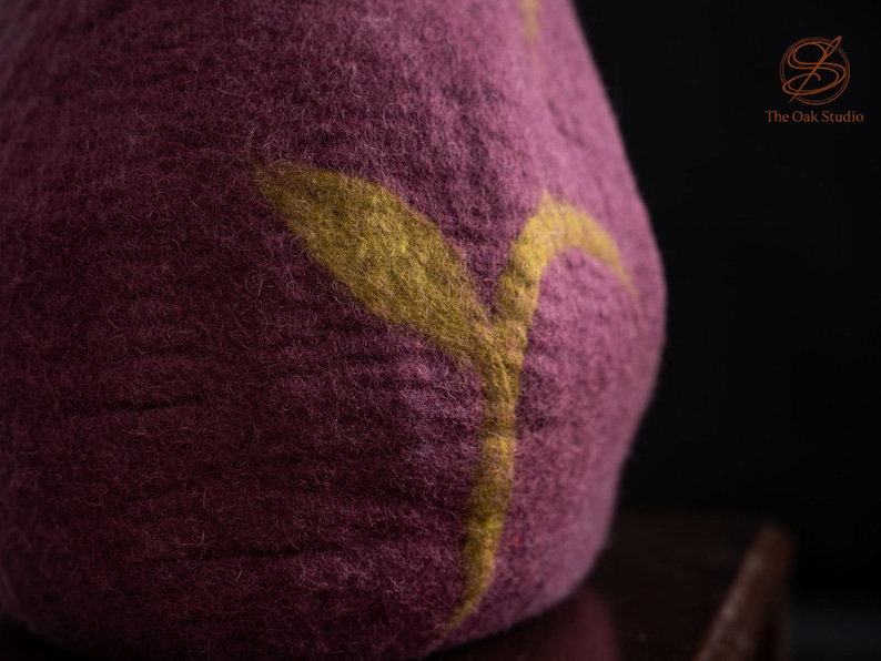 Peut inclure: Un objet en laine feutr&eacute;e magenta fonc&eacute; &agrave; la surface textur&eacute;e orn&eacute; d'un motif en forme de feuille jaune moutarde. Le coin sup&eacute;rieur droit affiche un logo avec le texte The Oak Studio en fine police dor&eacute;e sur fond sombre.