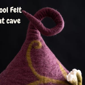 Peut inclure: Une grotte pour chat en laine feutr&eacute;e violet fonc&eacute; avec une pointe sup&eacute;rieure recourb&eacute;e. La surface pr&eacute;sente un motif d&eacute;coratif de vigne tourbillonnante vert olive et des accents floraux en feutre blanc. Le texte Wool Felt Cat cave est imprim&eacute;.