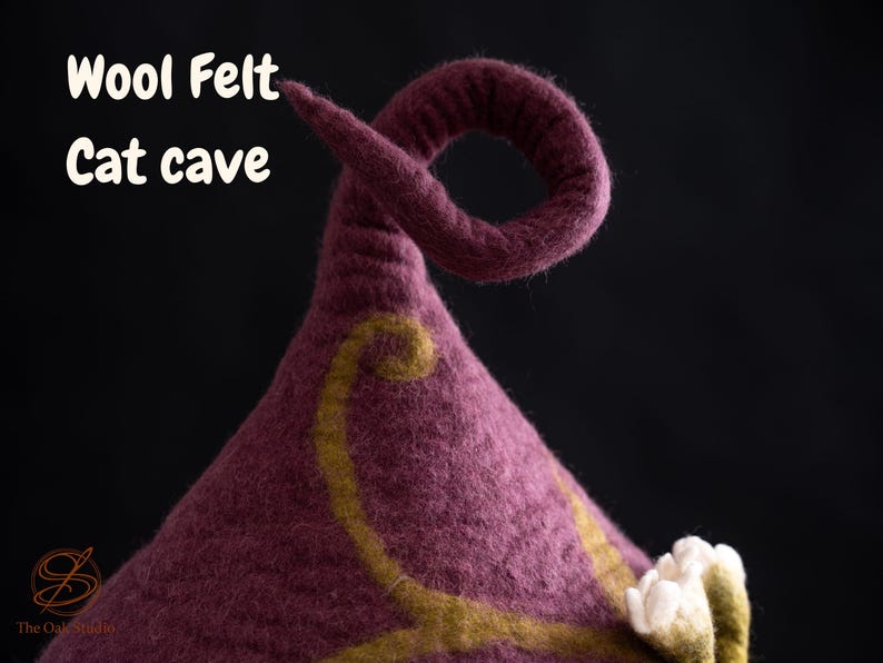 Peut inclure: Une grotte pour chat en laine feutr&eacute;e violet fonc&eacute; avec une pointe sup&eacute;rieure recourb&eacute;e. La surface pr&eacute;sente un motif d&eacute;coratif de vigne tourbillonnante vert olive et des accents floraux en feutre blanc. Le texte Wool Felt Cat cave est imprim&eacute;.