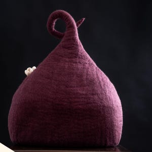 Peut inclure: Un chapeau en feutre de couleur prune profonde avec un sommet haut, pointu et recourb&eacute;, orn&eacute; d'une petite fleur de couleur claire sur le c&ocirc;t&eacute;. Le chapeau pr&eacute;sente une surface textur&eacute;e et dense en laine avec une forme conique distincte.