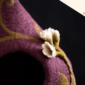 Peut inclure: Une structure en laine feutr&eacute;e couleur prune avec une ouverture arrondie, orn&eacute;e d'un motif de vigne vert olive et de petites fleurs blanches en feutre. Le coin sup&eacute;rieur droit affiche un logo avec le texte The Oak Studio en italique.