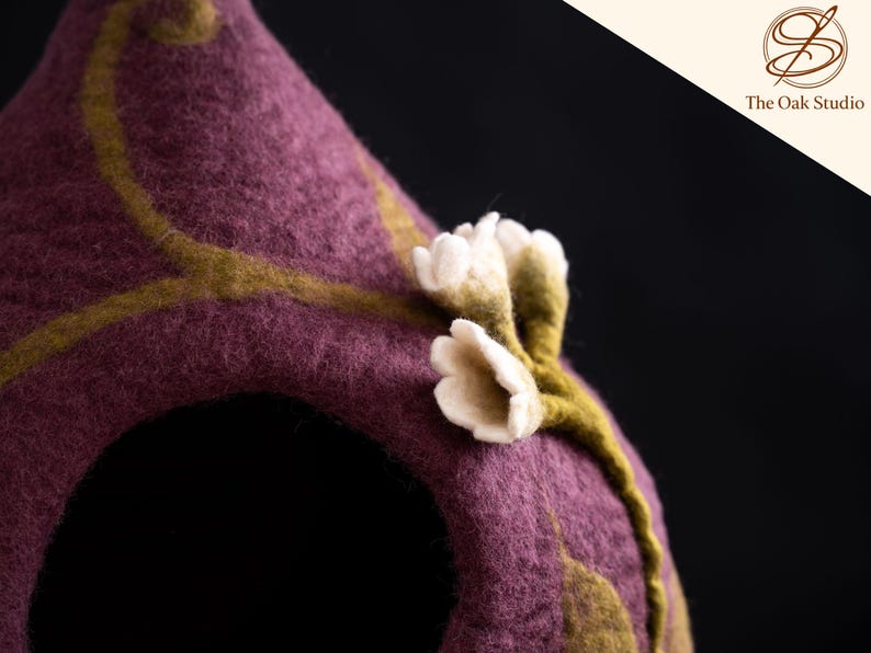 Peut inclure: Une structure en laine feutr&eacute;e couleur prune avec une ouverture arrondie, orn&eacute;e d'un motif de vigne vert olive et de petites fleurs blanches en feutre. Le coin sup&eacute;rieur droit affiche un logo avec le texte The Oak Studio en italique.