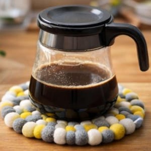 Könnte beinhalten: Eine Glaskaffeekaraffe mit schwarzem Deckel und Griff, gefüllt mit dunklem Kaffee, steht auf einem runden Filzuntersetzer. Der Untersetzer besteht aus grauen, gelben und weißen Filzkugeln und hat eine strukturierte Oberfläche.