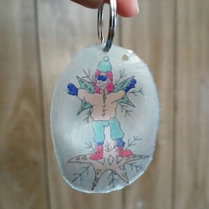 May include: A clear plastic keychain with a hand-drawn image of a person wearing a blue hat, blue gloves, and red boots standing on a star. The person is surrounded by snowflakes.
