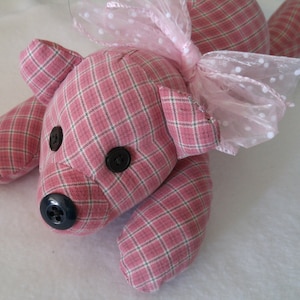 May include: A pink and white plaid teddy bear with black button eyes and a pink bow. The bear is lying on its side with its head turned to the right.
