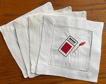 May include: Four white linen coasters with a decorative embroidered design. One coaster features a red matchbox with black outlines and a single lit match. The coasters have a stitched border.