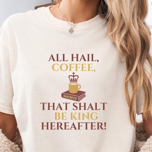 May include: Cream-colored t-shirt with the text "ALL HAIL, COFFEE, THAT SHALT BE KING HEREAFTER!" in brown and gold. A coffee cup with a crown sits atop a book labeled "Macbeth".