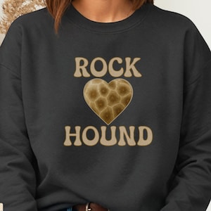 May include: Black sweatshirt with the text "ROCK HOUND" in a tan color, with a heart-shaped graphic in the center. The heart graphic has a textured, brown pattern. The sweatshirt is a crew neck style.