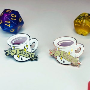 May include: Two enamel pins featuring a white coffee cup with a purple liquid inside. The cups have a gold rim and a gold handle. The text on the banner reads " +3 Energy".