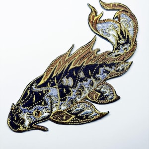 May include: An embroidered patch in the shape of a fish, featuring a dark navy blue body with intricate metallic gold and light blue thread detailing. The design includes flowing fins and a tail, with a textured finish created by dense stitching.