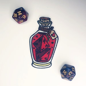 May include: An embroidered patch shaped like a glass bottle filled with red liquid and two dark purple twenty-sided dice. The bottle has a cork stopper, a brown rope tied around the neck, and a small tag with a red heart. Two purple dice sit nearby.