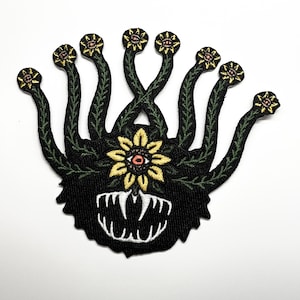 Könnte beinhalten: Gestickter Aufnäher mit einem stilisierten Monsterdesign. Der Aufnäher hat einen schwarzen Hintergrund mit einer zentralen Blume und mehreren Augenstielen. Die Blume ist gelb mit einem roten Zentrum, und die Augenstiele haben kleine blütenartige Details. Das Monster hat einen weißen Mund mit scharfen Zähnen.
