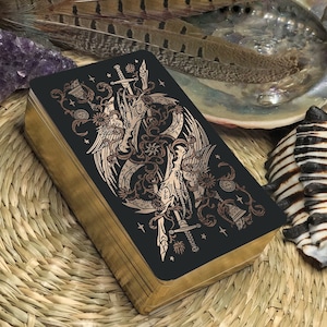 May include: A black tarot card with gold foil accents depicting two winged creatures intertwined with swords and vines. The card is surrounded by a wooden frame.