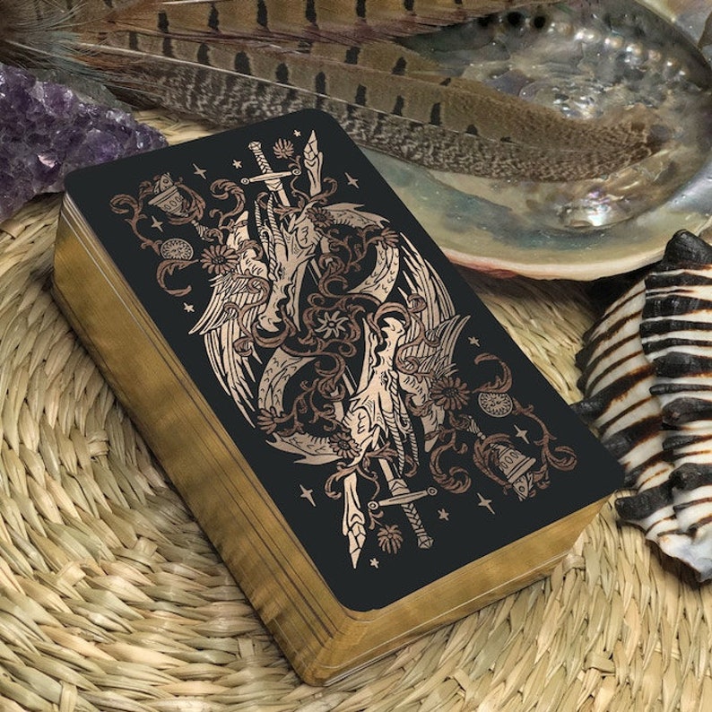 May include: A black tarot card with gold foil accents depicting two winged creatures intertwined with swords and vines. The card is surrounded by a wooden frame.