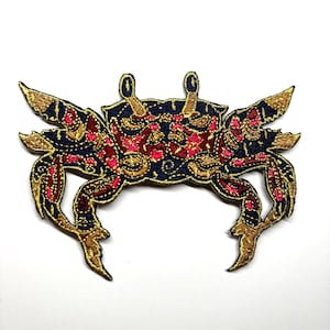 May include: An embroidered iron-on patch shaped like a crab. The design features a dark navy blue base with intricate metallic gold thread detailing and vibrant pink accents throughout the shell and legs, creating a textured, decorative appearance.