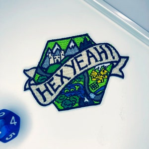 May include: A hexagonal embroidered patch featuring a landscape with a castle, mountains, and a forest in shades of green, blue, and white. A white banner across the center displays the black text "HEX YEAH!" alongside a blue multi-sided die.