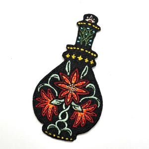 May include: Black embroidered patch in the shape of a decorative bottle. The patch features red and orange flowers with green vines and leaves. The bottle's neck has blue and green floral accents and yellow dots.